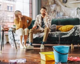 A couple in their living room, looking at a leak while attempting to book a plumber online.