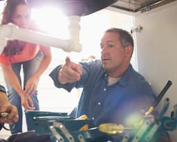 A rogue tradesperson gesturing under the sink, explaining a plumbing problem to a customer.